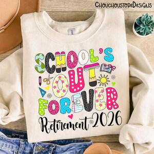 May include: Cream-colored sweatshirt with a colorful graphic that reads "SCHOOL'S OUT FOREVER Retirement 2026." The text features various patterns and colors, including pink polka dots, floral designs, and a pencil. The design includes a sun, scissors, and a book.