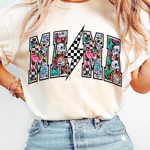 May include: Cream-colored t-shirt with the word "MAMA" in a colorful, patterned design. The letters are filled with skulls, flowers, and other graphics. A black and white checkered lightning bolt is in the center of the word.