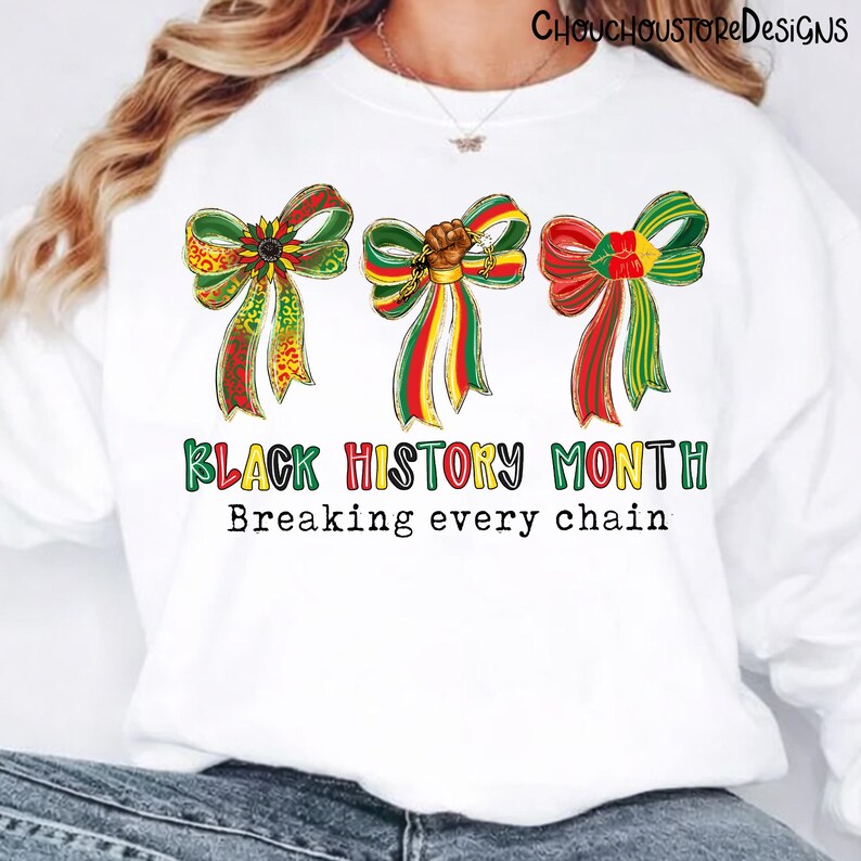 Op de afbeelding: Witte sweatshirt met drie decoratieve strikken in rood, groen en geel, met de tekst "Black History Month" en "Breaking every chain". De strikken hebben gedetailleerde patronen en een vuistontwerp.