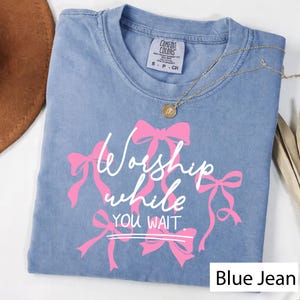 May include: A blue jean Comfort Colors t-shirt with the text "Worship while you wait" in white script and pink bows. A gold necklace with a pendant is draped on the shirt.