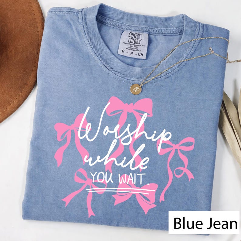 May include: A blue jean Comfort Colors t-shirt with the text "Worship while you wait" in white script and pink bows. A gold necklace with a pendant is draped on the shirt.
