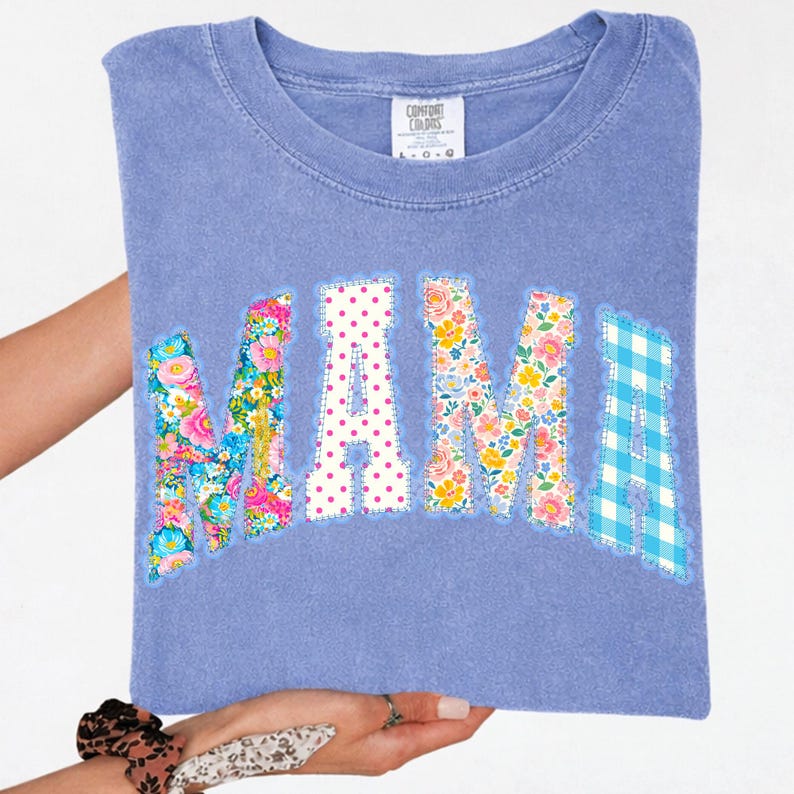 May include: A periwinkle blue t-shirt with the word "MAMA" appliqu&eacute;d across the front. The letters are decorated with floral patterns, polka dots, and a blue gingham check. The shirt is held up by a person.