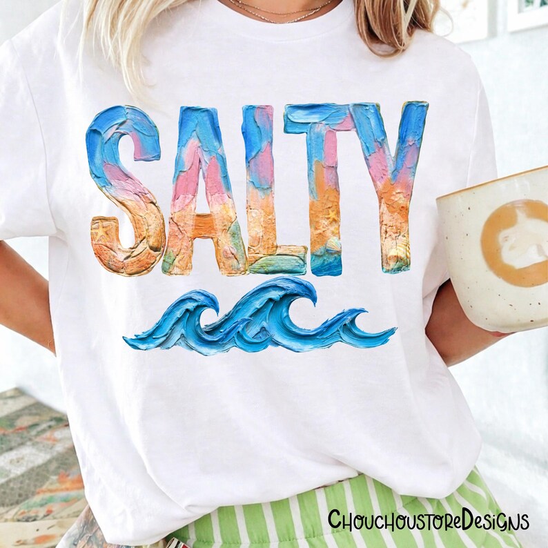 May include: White t-shirt with the word "SALTY" in colorful, textured lettering, and blue wave graphics. The shirt also features the text "ChouchoustoreDesigns" at the bottom. A person is holding a mug.