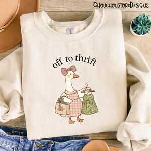May include: A cream-coloured sweatshirt featuring a cartoon goose with a bow and apron, carrying a handbag and a dress. The words "off to thrift" are above the goose. The design uses soft colours and has a whimsical feel.