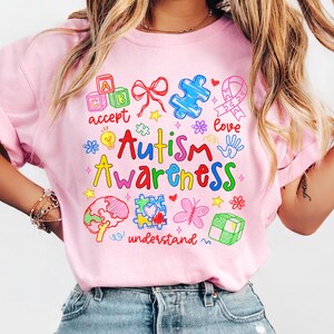 May include: A light pink t-shirt with the words "Autism Awareness" in colorful lettering. The shirt features various graphics, including puzzle pieces, a ribbon, a brain, and the words "accept", "love", and "understand".