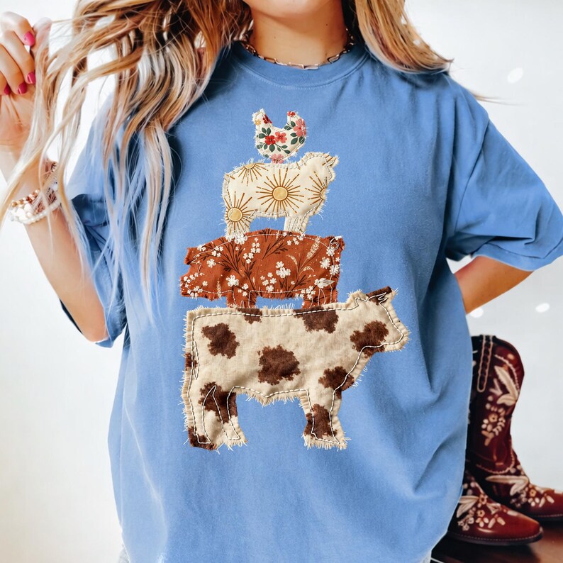 May include: A light blue t-shirt featuring a stacked farm animal design. The design includes a cow, pig, sheep, and chicken, each with a unique fabric pattern. The cow is at the bottom, with brown spots, and the chicken at the top, with floral accents.