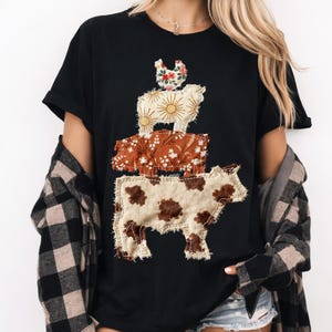 May include: Black t-shirt featuring a stacked animal design. The design includes a cow with brown spots, a pig with a floral pattern, a sheep with sunburst details, and a chicken with a floral pattern. The shirt has short sleeves and a crew neck.