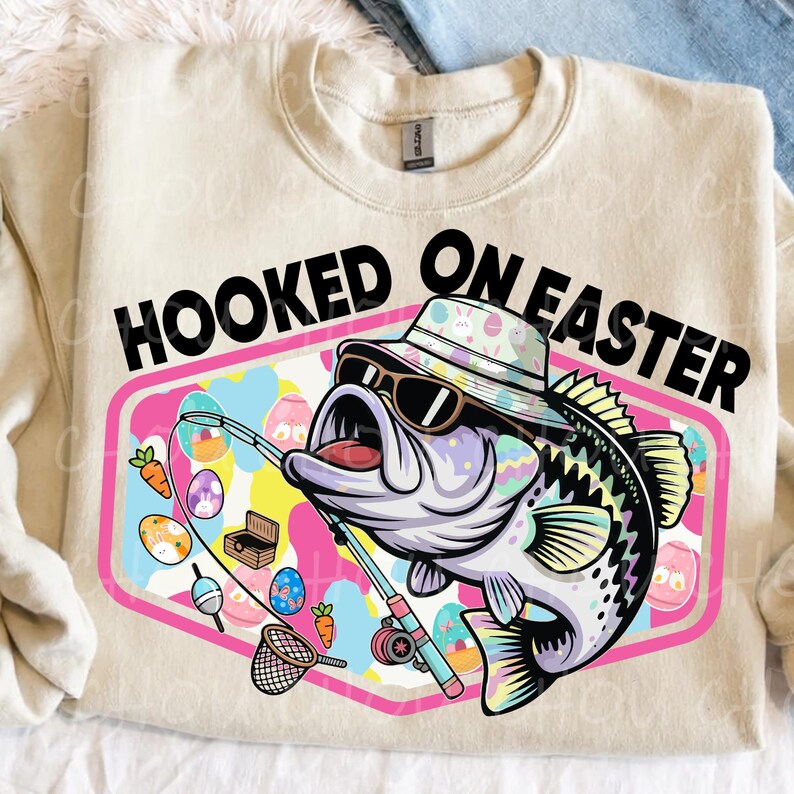 Pode incluir: Camisola bege com a frase "HOOKED ON EASTER". O gr&aacute;fico apresenta um baixo de desenho animado usando &oacute;culos de sol e um chap&eacute;u de pescador, segurando uma vara de pescar com ovos de P&aacute;scoa e cenouras. O design &eacute; emoldurado em um hex&aacute;gono rosa.
