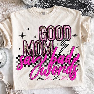 May include: Cream-colored t-shirt with the text "GOOD MOM say bad words" in pink and leopard print. The design includes a checkered pattern, lightning bolt, and star accents. The shirt has a crew neck and short sleeves.