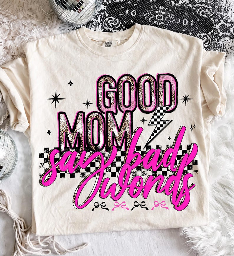 May include: Cream-colored t-shirt with the text "GOOD MOM say bad words" in pink and leopard print. The design includes a checkered pattern, lightning bolt, and star accents. The shirt has a crew neck and short sleeves.