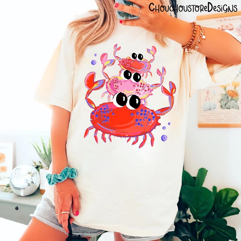 May include: Cream-colored t-shirt featuring a whimsical illustration of three cartoon crabs stacked on top of each other. The crabs are in shades of red and pink with large, expressive eyes and purple accents. The text "CHOUCHOUSTOREDESIGNS" is at the top.