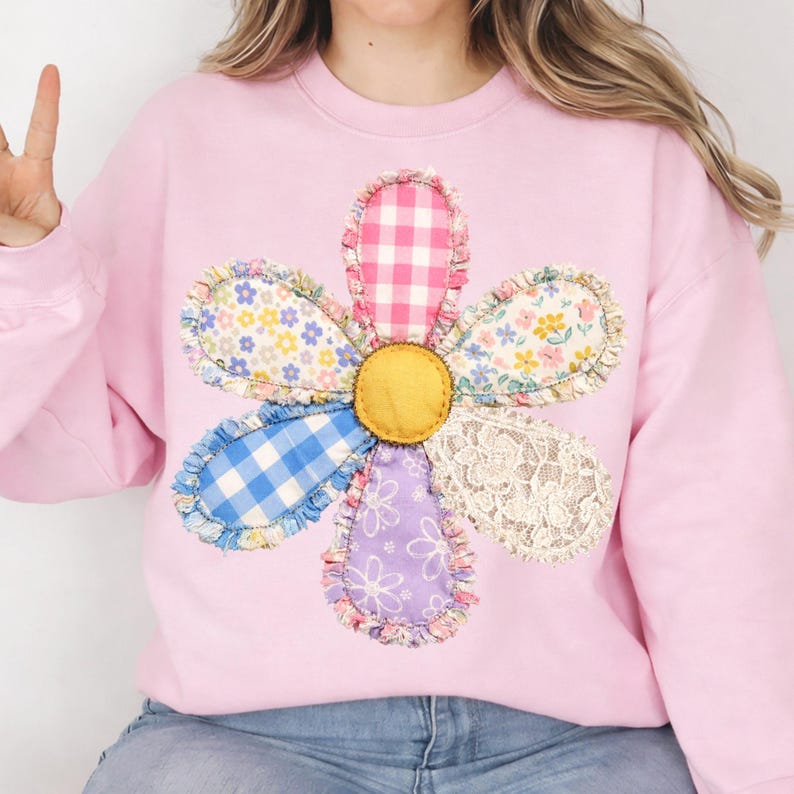 May include: A light pink sweatshirt featuring a large, appliqued flower design. The flower has a yellow center and petals made from various patterned fabrics, including gingham, floral, and lace. The sweatshirt is a casual, comfortable style.