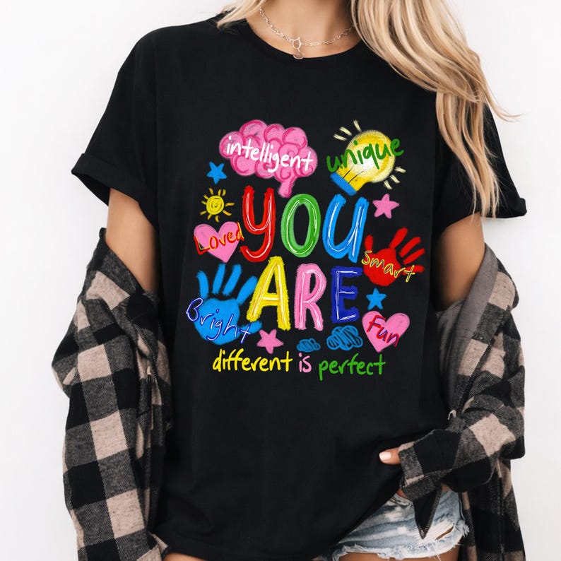 May include: Black t-shirt featuring a vibrant graphic with the words "YOU ARE" in bold letters, encircled by words such as "intelligent," "unique," "loved," "bright," "smart," and "fun." The phrase "different is perfect" is at the bottom.