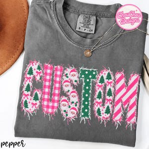 Könnte beinhalten: Ein anthrazitfarbenes Kurzarm-T-Shirt mit dem Wort CUSTOM in großen Applikationsbuchstaben mit ausgefransten Rändern. Jeder Buchstabe zeigt ein anderes Muster, darunter rosa Vichy-Karo, grüne Punkte, Weihnachtsmänner und Tannen.