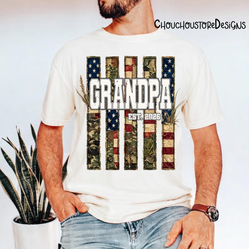May include: Cream-colored t-shirt featuring a patriotic design with the word "GRANDPA" in bold white letters. The design incorporates elements of the American flag and camouflage patterns, with the text "EST. 2026" below. The shirt is a casual wear item.