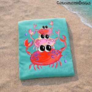 May include: A turquoise beach towel featuring a stack of three cartoon crabs in shades of red and pink. The crabs have large, expressive eyes and are decorated with blue dots. The towel is on a sandy surface.
