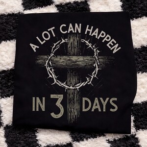 Puede incluir: Camiseta negra con un gr&aacute;fico desgastado que presenta una cruz, una corona de espinas y el texto "A LOT CAN HAPPEN IN 3 DAYS" en blanco y gris. El dise&ntilde;o tiene un aspecto vintage y desgastado.