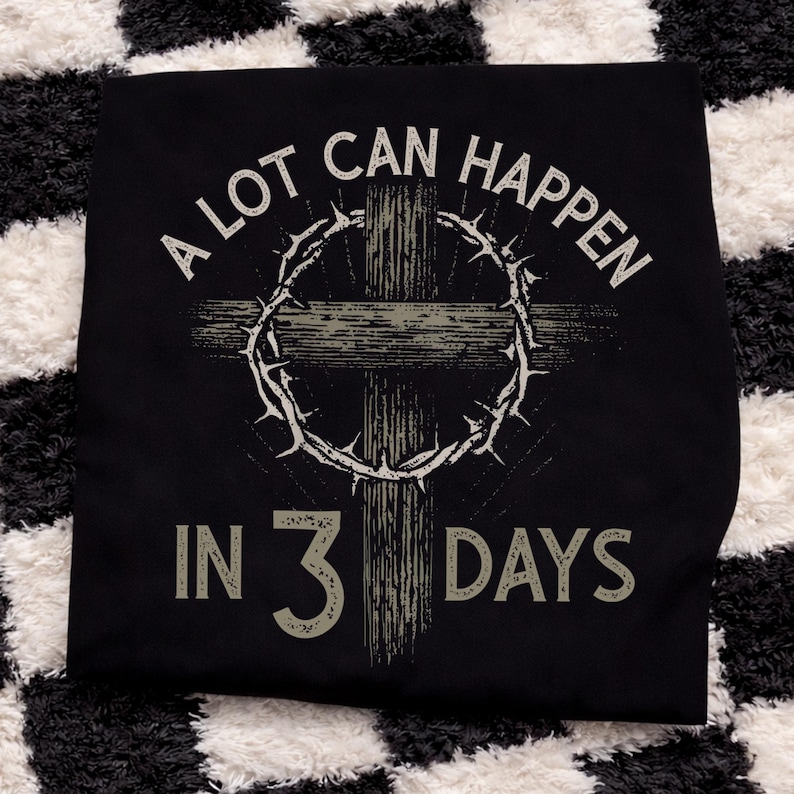 Puede incluir: Camiseta negra con un gr&aacute;fico desgastado que presenta una cruz, una corona de espinas y el texto "A LOT CAN HAPPEN IN 3 DAYS" en blanco y gris. El dise&ntilde;o tiene un aspecto vintage y desgastado.