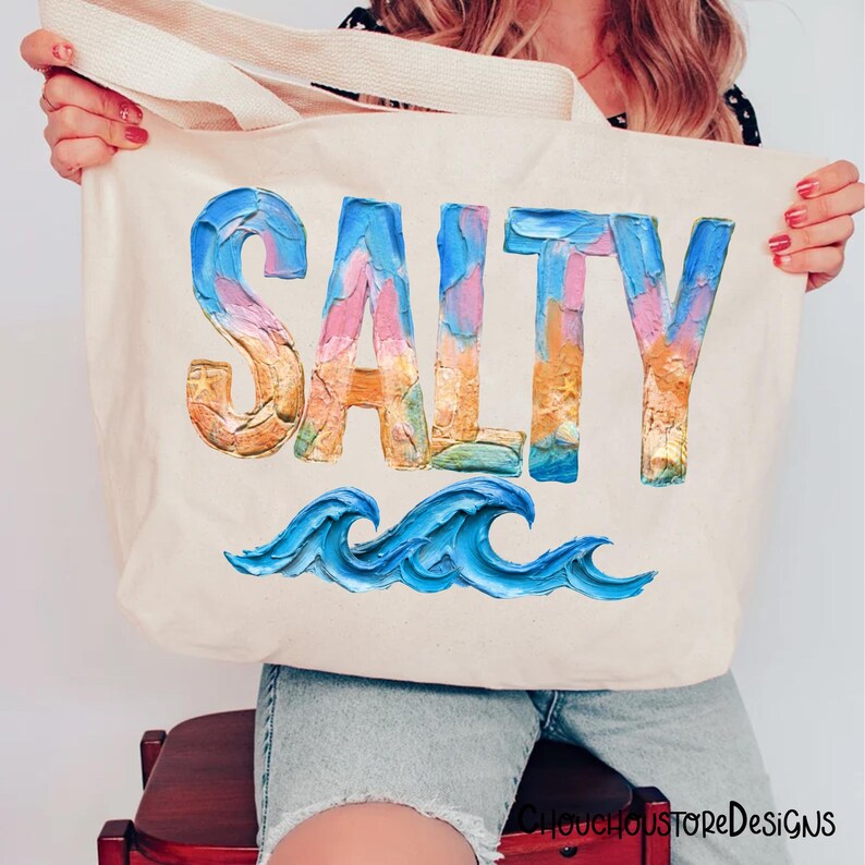 May include: A cream-colored tote bag with the word "SALTY" in large, colorful, textured letters. Below the word are blue painted waves. The bag has a simple design and is held up by the handles.