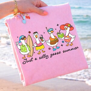 May include: Pink beach towel featuring a cartoon illustration of four geese enjoying summer activities. The geese are wearing sunglasses, holding drinks, and a surfboard. Text reads "Just a silly goose summer."