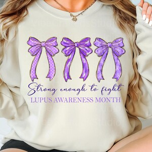 K&ouml;nnte beinhalten: Cremefarbenes Sweatshirt mit drei lila Schleifen mit goldenen Verzierungen. Die Schleifen haben wei&szlig;e Tupfen und lila B&auml;nder. Der Text lautet "Strong enough to fight" und "LUPUS AWARENESS MONTH".