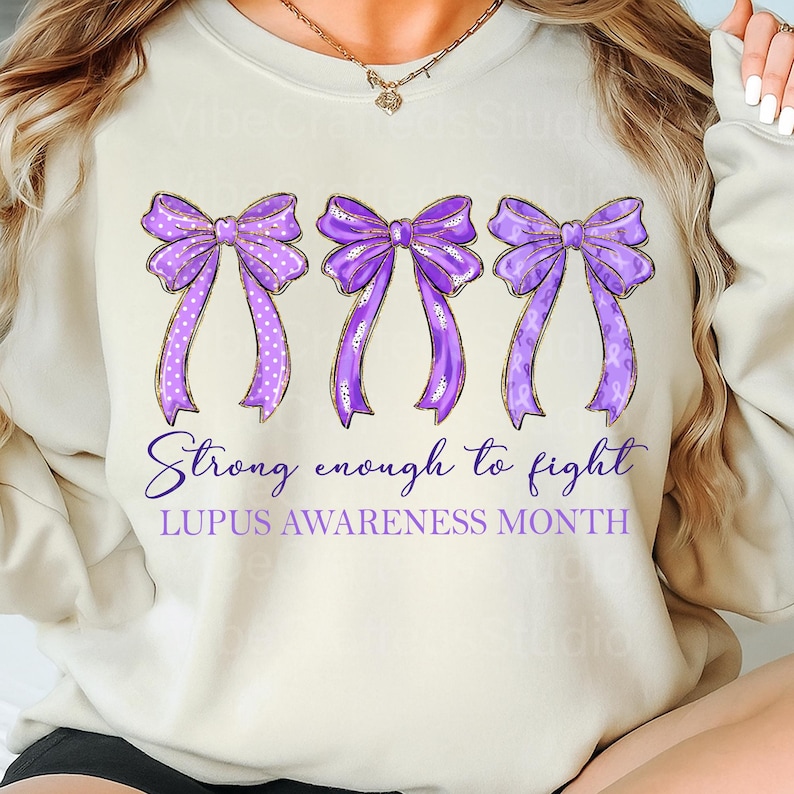 K&ouml;nnte beinhalten: Cremefarbenes Sweatshirt mit drei lila Schleifen mit goldenen Verzierungen. Die Schleifen haben wei&szlig;e Tupfen und lila B&auml;nder. Der Text lautet "Strong enough to fight" und "LUPUS AWARENESS MONTH".