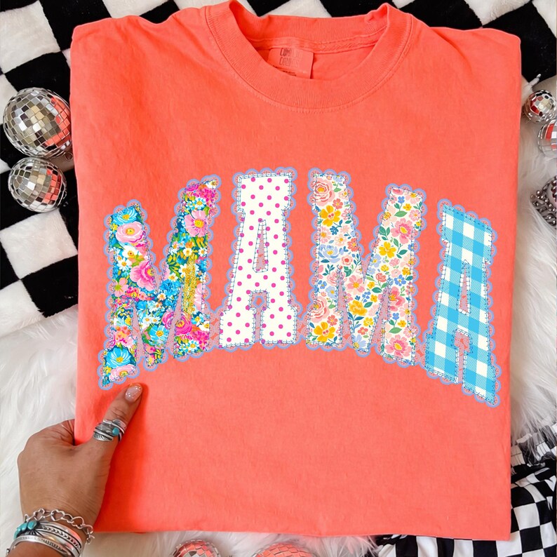 May include: Coral-coloured t-shirt featuring the word "MAMA" in patchwork letters. The letters are decorated with floral, polka dot, and gingham patterns. The shirt has a crew neck.