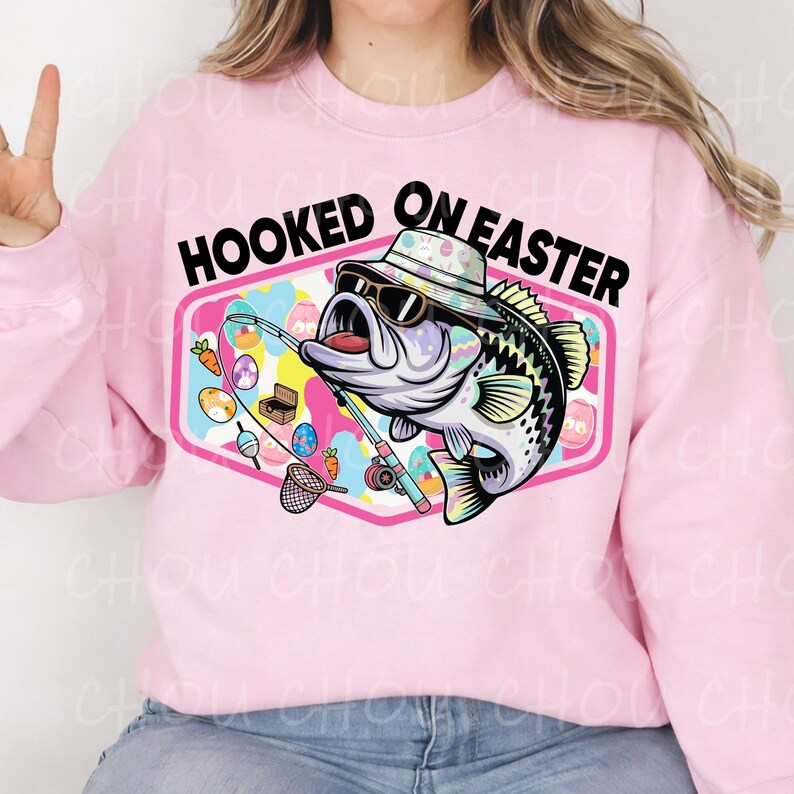 Pode incluir: Camisola rosa com o texto "HOOKED ON EASTER" acima de um gr&aacute;fico de um peixe usando &oacute;culos de sol e um chap&eacute;u, segurando uma vara de pesca. O gr&aacute;fico inclui ovos de P&aacute;scoa e uma rede de pesca.