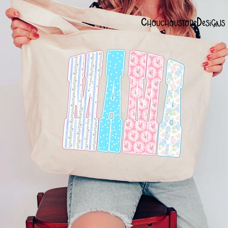 May include: Beige tote bag with the word "MAMA" in large, colorful letters. The letters are decorated with floral and polka dot patterns in blue, pink, and white. The bag has a simple design and is held by a person.