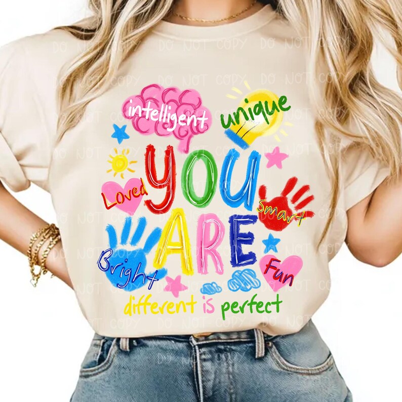 May include: A cream-coloured t-shirt featuring a vibrant graphic. The design prominently displays the words "YOU ARE" in various colours, accompanied by words such as "intelligent", "unique", "loved", "bright", and "fun". The phrase "different is perfect" is also included.
