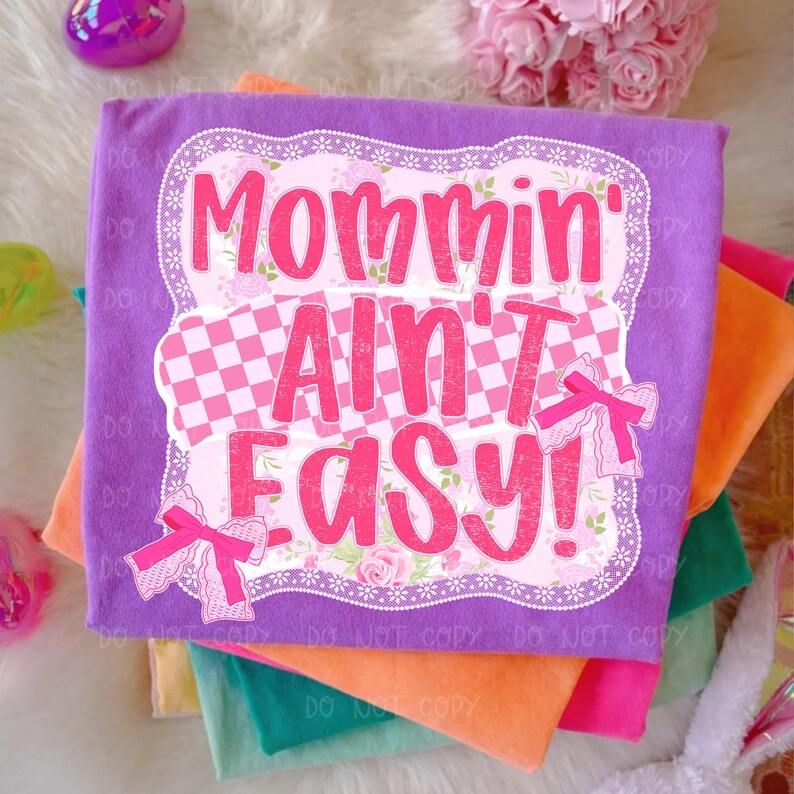 May include: A stack of colorful t-shirts, with a purple shirt on top. The top shirt has the phrase "Mommin' Ain't Easy!" in pink, with a floral and checkered design. Pink bows are on the shirt.