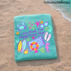May include: A turquoise beach towel with colorful illustrations of summer items, including a crab, sunglasses, a life preserver, and the text "I Was Made For Happy Sunny Days". The towel is lying on a sandy beach.