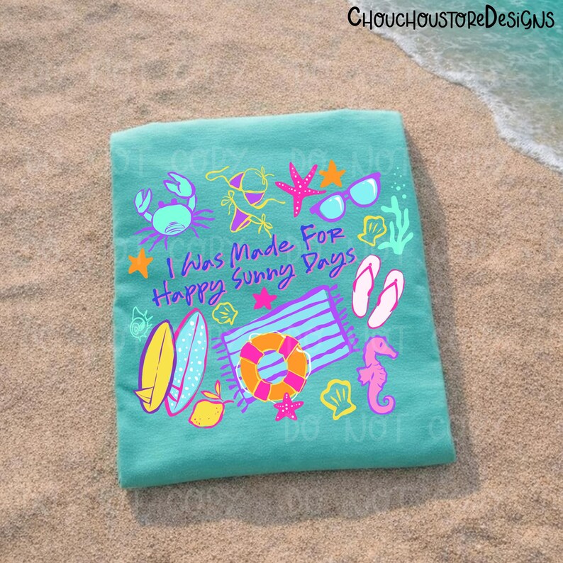 May include: A turquoise beach towel with colorful illustrations of summer items, including a crab, sunglasses, a life preserver, and the text "I Was Made For Happy Sunny Days". The towel is lying on a sandy beach.