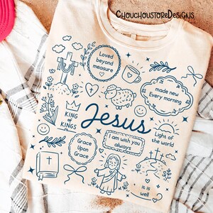 May include: Cream-colored sweatshirt with a navy blue graphic design featuring the word "Jesus" and various religious symbols and phrases, including a cross, heart, and sheep. The text "CHOUCHOUSTORE DESIGNS" is at the top.