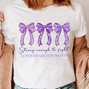 K&ouml;nnte beinhalten: Wei&szlig;es T-Shirt mit drei lila Schleifen, goldenen Umrissen und wei&szlig;en Punkten. Der Text lautet "Strong enough to fight" und "Lupus Awareness Month". Ein l&auml;ssiges T-Shirt f&uuml;r das Bewusstsein.
