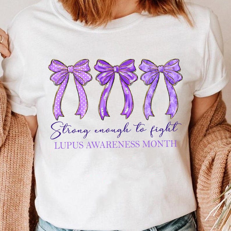 K&ouml;nnte beinhalten: Wei&szlig;es T-Shirt mit drei lila Schleifen, goldenen Umrissen und wei&szlig;en Punkten. Der Text lautet "Strong enough to fight" und "Lupus Awareness Month". Ein l&auml;ssiges T-Shirt f&uuml;r das Bewusstsein.
