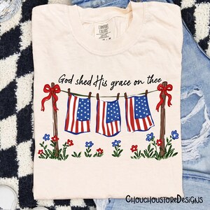 May include: Cream-colored t-shirt featuring a patriotic design. The graphic includes American flags hanging on a clothesline, red bows, and the text "God shed His grace on thee." Floral accents are at the bottom.