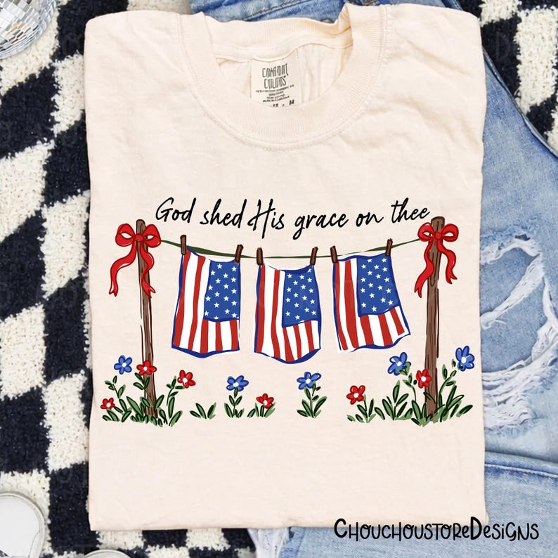 May include: Cream-colored t-shirt featuring a patriotic design. The graphic includes American flags hanging on a clothesline, red bows, and the text "God shed His grace on thee." Floral accents are at the bottom.