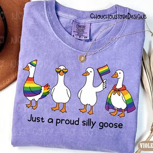 K&ouml;nnte beinhalten: Ein lila Comfort Colors T-Shirt mit vier Cartoon-G&auml;nsen, die Regenbogen-Pride-Accessoires tragen. Der Text "Just a proud silly goose" ist darunter aufgedruckt. Eine goldene Halskette liegt &uuml;ber dem Shirt.