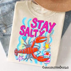 Puede incluir: Camiseta color crema con un dise&ntilde;o gr&aacute;fico vibrante. El dise&ntilde;o presenta las palabras "STAY SALTY" en rosa brillante, una langosta roja, una lata de cerveza y otros elementos n&aacute;uticos. La camiseta es una prenda casual.