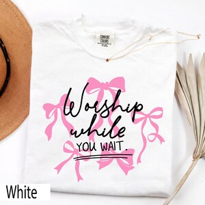 May include: White t-shirt with the text "Worship while you wait" in black script and pink bow graphics. The shirt has a crew neck and a tag that says "Comfort Colors". A gold necklace with a star pendant is draped on the shirt.