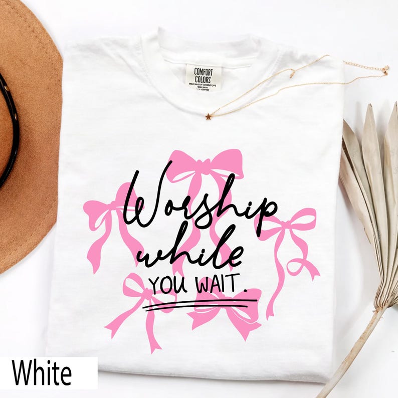 May include: White t-shirt with the text "Worship while you wait" in black script and pink bow graphics. The shirt has a crew neck and a tag that says "Comfort Colors". A gold necklace with a star pendant is draped on the shirt.