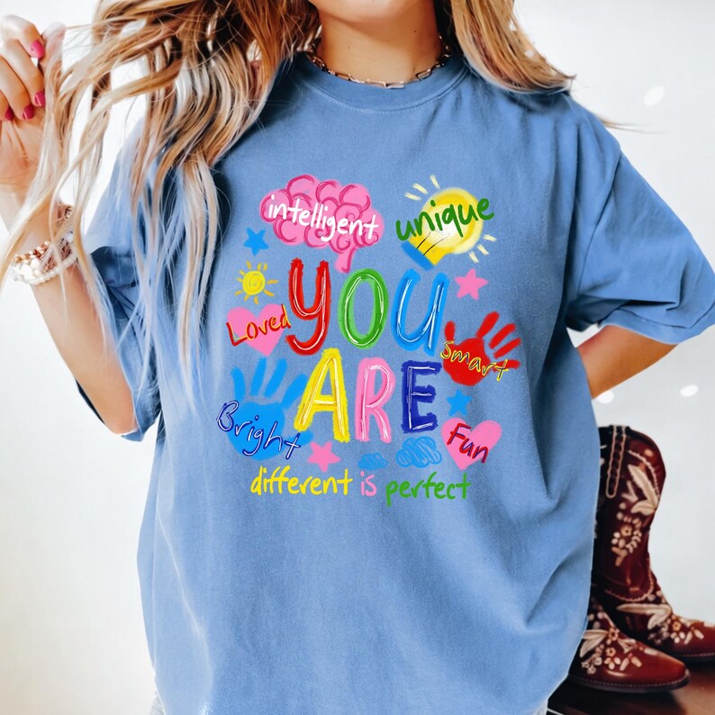 May include: A pale blue t-shirt featuring a vibrant graphic with the words "YOU ARE" in various colours, surrounded by words such as "intelligent", "unique", "loved", "bright", "smart", and "fun". The phrase "different is perfect" is also included.