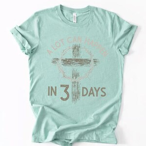 Puede incluir: Camiseta verde claro con el texto "A LOT CAN HAPPEN IN 3 DAYS". El dise&ntilde;o incluye una cruz y una corona de espinas en tonos rosa y marr&oacute;n apagados. El texto est&aacute; en una fuente desgastada.
