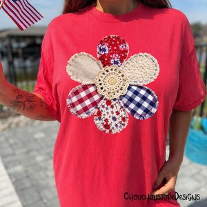 May include: A red t-shirt with a large, appliqued flower design. The flower has petals in various patterns: red floral, gingham, and blue gingham. The centre is a cream-coloured crochet doily. The text "ChouchoustoreDesigns" is at the bottom.