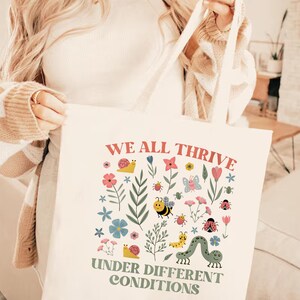 Peut inclure: Sac fourre-tout cr&egrave;me avec le texte "WE ALL THRIVE UNDER DIFFERENT CONDITIONS" en corail et vert olive. Le sac est orn&eacute; d'illustrations color&eacute;es de fleurs, d'insectes et de feuillages.