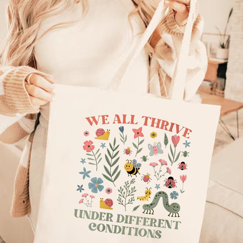 Peut inclure: Sac fourre-tout cr&egrave;me avec le texte "WE ALL THRIVE UNDER DIFFERENT CONDITIONS" en corail et vert olive. Le sac est orn&eacute; d'illustrations color&eacute;es de fleurs, d'insectes et de feuillages.