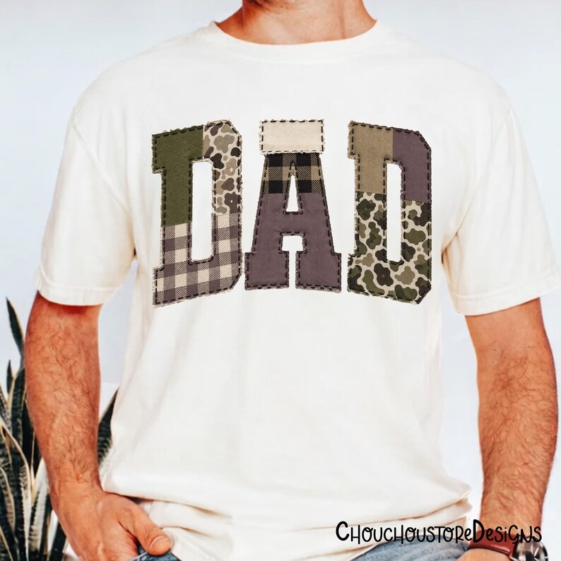 May include: Cream-colored t-shirt with the word "DAD" in a patchwork design. The letters feature various patterns, including plaid and camouflage, stitched with dark thread. The shirt is a casual style.