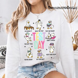 May include: White sweatshirt with a colorful graphic featuring cartoon ducks in various poses with text like "Test Day Ready" and "You Got This." The word "TEST DAY" is prominently displayed in the center.