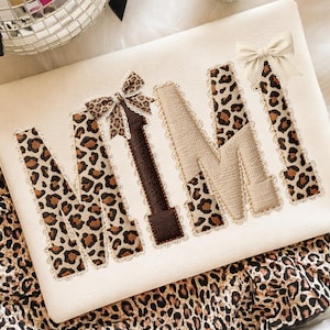 May include: Cream-colored decorative pillow with the word "MIMI" embroidered in a mix of leopard print, brown, and beige fabrics. Each letter is outlined with a delicate lace trim, and small bows adorn the "M" and "I".